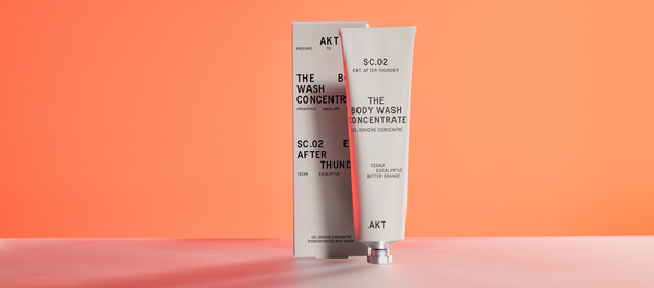 A tube of AKT Body Wash Concentrate SC.02 in After Thunder scent, positioned in front of its silver packaging box against an orange background.